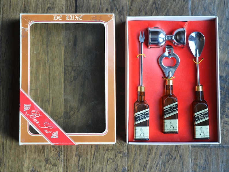 May include: A red and gold box with a clear window showing a bar set. The set includes three miniature bottles, a bottle opener, a jigger, and a cocktail spoon. The box says "De Luxe Bar Set" on the front.