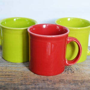 Op de afbeelding: Drie keramische mokken, één rood, twee groen, op een houten oppervlak.