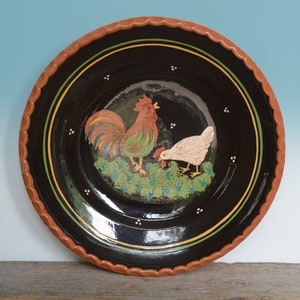 Op de afbeelding: Een zwarte keramische plaat met een bruine rand en een handgeschilderd ontwerp van een haan en een hen. De haan is rood, groen en geel, en de hen is wit. De plaat heeft een zwarte achtergrond met witte stippen.