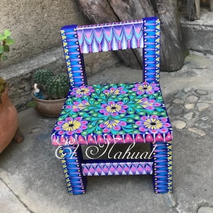 May include: A small, hand-painted wooden chair with a blue base and a colorful floral design on the seat. The chair is decorated with pink, yellow, green, and blue paint. The chair is a traditional Mexican folk art piece.