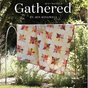 Könnte beinhalten: Ein Quilt mit Sternmuster in Rot-, Gelb- und Brauntönen hängt an einem Metallgestell. Im Hintergrund ist das Buch "Gathered" von Jen Kingwell zu sehen. Der Quilt wird im Freien präsentiert, was auf ein handgefertigtes Produkt hindeutet.