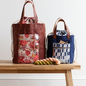 May include: Two canvas tote bags with leather handles and drawstring closures. The larger bag is brown with a floral print and a zippered pocket. The smaller bag is navy blue with a geometric print and a zippered pocket. Both bags are sitting on a wooden bench with yarn balls in front of them.