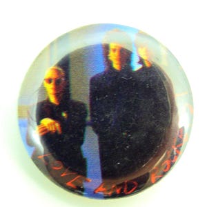 May include: A round, clear button with a blurred image of three people in dark clothing. The button has the words "LOVE AND ROCKETS" in red text around the edge. The background is white.