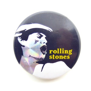 May include: A black and white button with a picture of a person singing. The text "rolling stones" is in yellow letters.