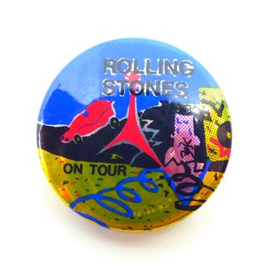 May include: A round button with a blue and yellow background featuring the text "ROLLING STONES ON TOUR." The button has a red car, a red tower, and other colorful graphics.