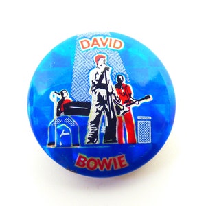 May include: A blue and white button with a picture of David Bowie on stage with a band. The button says "DAVID BOWIE" in red letters.