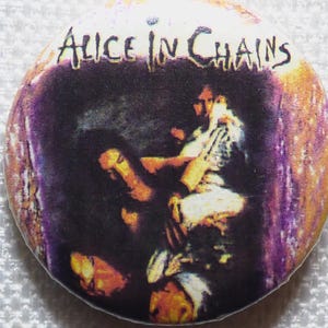 Pode incluir: Botão redondo com o texto "Alice in Chains" em branco, sobre uma imagem escura de figuras. O botão tem uma borda roxa e dourada, e é provavelmente um item de colecionador ou merchandising.