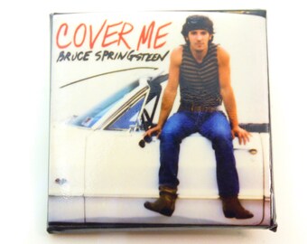 May include: A vintage album cover featuring Bruce Springsteen, with the title "Cover Me" in red lettering. The image shows Springsteen sitting on the back of a white car, wearing a striped tank top and blue jeans.