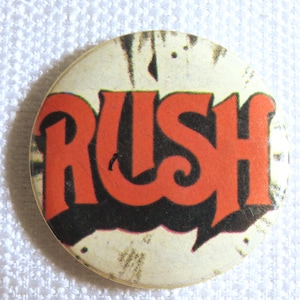 May include: A circular badge featuring the word "RUSH" in red, decorative lettering. The letters are outlined in black, set against a cream-coloured background. The badge has a vintage, slightly worn appearance.
