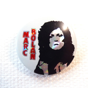 Könnte beinhalten: Ein weißer Knopf mit einem Schwarzweißbild von Marc Bolan. Der Text "MARC BOLAN" ist in roten Buchstaben auf dem Knopf gedruckt.