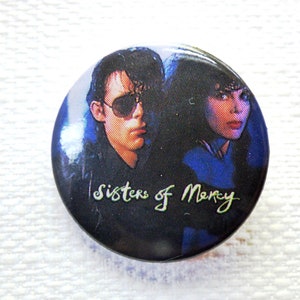 May include: A black and white button badge featuring a photo of two people with the text "Sisters of Mercy" in green cursive.