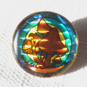 Peut inclure: Épinglette ronde avec un motif holographique de champignons bruns et d'une fleur jaune. L'épinglette a une bordure dorée et un fond vert d'eau et jaune. Le motif est sur un fond blanc.