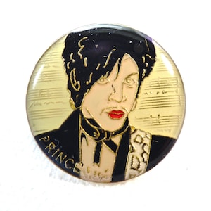 May include: A round, gold-colored pin with a black and white image of Prince. The background is a light yellow with gold musical notes. The pin says "Prince" in gold letters.