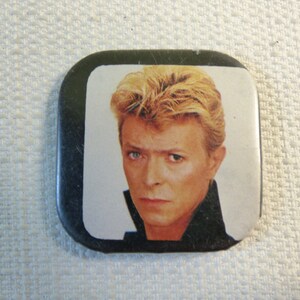 Puede incluir: Un botón cuadrado con un borde negro y un fondo blanco. El botón presenta una fotografía en color de David Bowie con el pelo rubio y una chaqueta negra.