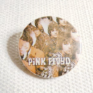 May include: A round button with a photo of the band Pink Floyd. The button features a close-up of the band members, with the band's name printed in white letters on a brown background.