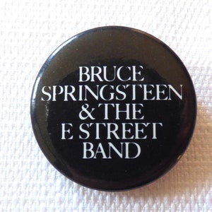 May include: Black and white button with the text "Bruce Springsteen & The E Street Band" in white lettering.