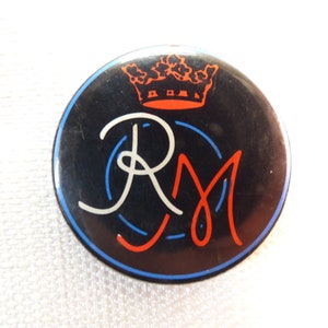Op de afbeelding: Een zwarte en witte knop met een rode en blauwe kroon en de letters "RM" in wit met rode en blauwe contouren.
