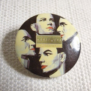 Puede incluir: Un botón redondo con una imagen en blanco y negro de cuatro caras. El texto "Kraftwerk" y "Electric Cafe" está impreso en amarillo en el botón.