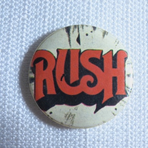 May include: A round, white button with a black and red graphic of the word "RUSH" in a stylized font. The background of the button has a black and white splatter pattern.