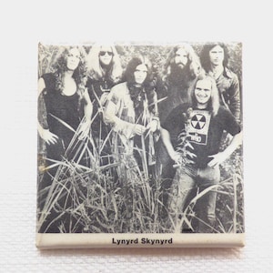 May include: Black and white photo of the band Lynyrd Skynyrd. The band members are standing in a field of tall grass. One band member is wearing a t-shirt with the words "The Who" on it.