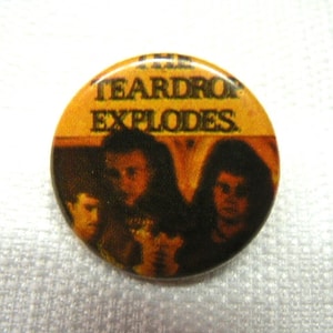 Può includere: Un bottone rotondo con sfondo giallo e testo nero che recita "The Teardrop Explodes". Il bottone presenta una foto in bianco e nero di una band con tre membri.