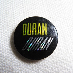 Op de afbeelding: Een zwarte knop met een zilveren en geel holografisch ontwerp met de tekst "Duran Duran".
