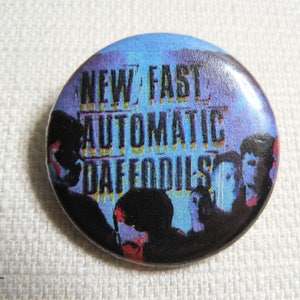 Puede incluir: Un botón azul y blanco con el texto "New Fast Automatic Daffodils" en letras negras y amarillas. El botón presenta una imagen en blanco y negro de una banda.
