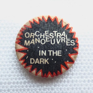 Może przedstawiać: Okrągła przypinka z czarnym środkiem i poszarpaną czerwoną obwódką. Napis "ORCHESTRAL MANOEUVRES IN THE DARK" jest nadrukowany białymi wielkimi literami na środku przypinki, otoczony jasnobeżowym tłem.