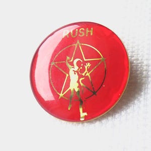 May include: A circular red pin featuring a gold-colored graphic of a person standing inside a star. The word RUSH is printed in gold letters at the top of the pin. The design has a glossy finish and a thin gold-colored metal border around the edge.
