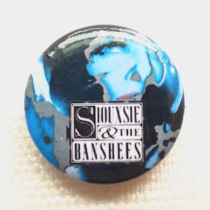 May include: A circular pinback button featuring an abstract blue, black, and white marbled pattern. Centered on the button is a rectangular white box containing the black text Siouxsie & The Banshees in a stylized serif font.