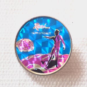 May include: A silver pin with a blue and pink holographic design. The pin features a figure standing on a wave with the text "Rush Hemispheres" above.