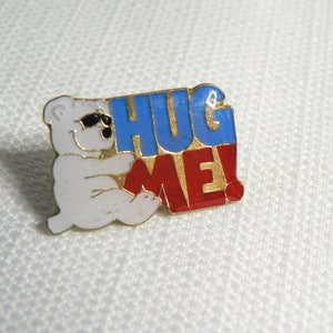 May include: A white polar bear pin wearing sunglasses, hugging a sign that says "HUG ME!" in blue and red letters with gold trim. The pin is a fun accessory.