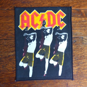 May include: Black and white patch with the band name "AC/DC" in red and yellow letters. Three figures in black and white are shown playing guitars.