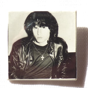 Puede incluir: Foto en blanco y negro de una persona con el pelo largo y oscuro que lleva una chaqueta de cuero negra y un collar con una cruz de plata.