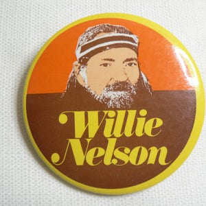 May include: A yellow and brown round button with a picture of Willie Nelson wearing a white headband and a brown beard. The text "Willie Nelson" is written in yellow letters on the button.