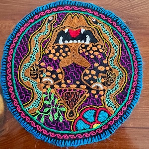 Op de afbeelding: Geborduurde ronde patch met een gedetailleerd ontwerp. De patch heeft een centraal beeld van een brullend dier met slangen, omgeven door een zwarte achtergrond met oranje en paarse accenten. De rand is blauw met roze en groene stiksels.