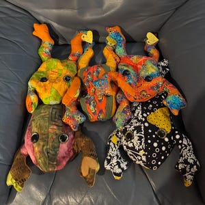 May include: A group of handmade frog plush toys in assorted colours and patterns. Each frog has large black button eyes and is made from patchwork fabric in shades of orange, green, pink, and black. Some have small yellow heart-shaped tags.