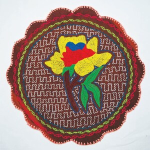 May include: A round, brown fabric coaster with a colorful embroidered flower design. The flower is yellow with red, blue, and green accents. The coaster is surrounded by a decorative border.