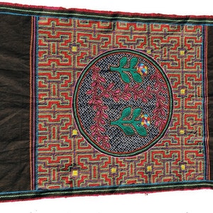May include: A black fabric textile with a colorful embroidered design. The design features a circular pattern with two stylized floral motifs in the center, surrounded by a geometric pattern.
