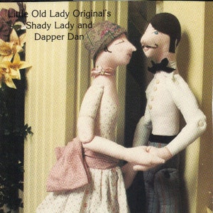 May include: Two fabric dolls, a woman and a man, are standing next to each other. The woman is wearing a white dress with pink accents and a floral pattern. The man is wearing a white shirt with a black bow tie and gray pants with a red stripe. The dolls are holding hands. The text "Little Old Lady Original's Shady Lady and Dapper Dan" is printed above the dolls.