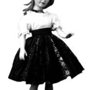 May include: A black and white photo of a doll wearing a white blouse with a black lace skirt and a black belt.