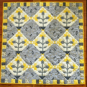 May include: A quilt with a geometric pattern of yellow and gray squares. The quilt features a repeating design of stylized trees with branches and leaves.