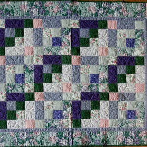 May include: A quilt with a patchwork design featuring squares of various floral prints in shades of pink, blue, green, and purple. The quilt is edged with a green floral border.