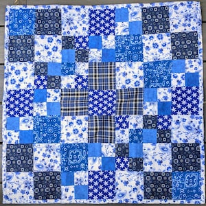 May include: A square patchwork quilt featuring a variety of blue and white fabric squares. The design includes patterns of flowers, paisley, geometric shapes, and plaid, arranged in a grid. The quilt is finished with a solid blue fabric border.