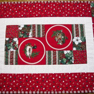 May include: Red and green Christmas table runner with a white border. The runner features a patchwork design with a duck and a squirrel in the center.