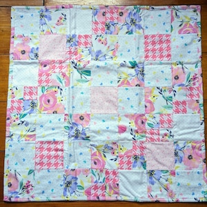 May include: A white baby quilt with a patchwork design featuring pink, blue, and white floral patterns. The quilt is made of squares and rectangles, and the seams are visible.