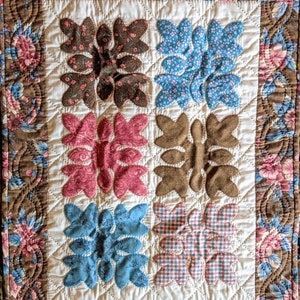 May include: A brown, white, and blue patchwork quilt with a floral pattern on the border. The quilt is made up of six squares, each with a different floral design. The quilt is quilted with a simple stitch.