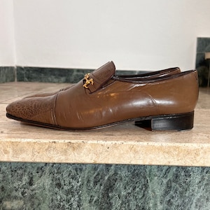 Könnte beinhalten: Braune Lederschuhe zum Hineinschlüpfen mit einem goldenen Kettendetail. Die Schuhe haben eine spitze Zehenpartie und einen niedrigen Absatz.