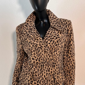 May include: A brown and black leopard print double-breasted jacket with gold button accents. The jacket is worn on a black mannequin with a gold and red patterned hat.