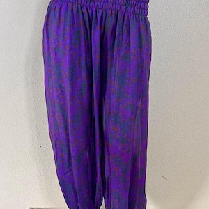 May include: Purple harem pants featuring a dark green and red floral pattern. These pants have a wide, smocked elastic waistband and elasticized cuffs at the ankles, creating a loose, flowing silhouette with a gathered, voluminous leg design.